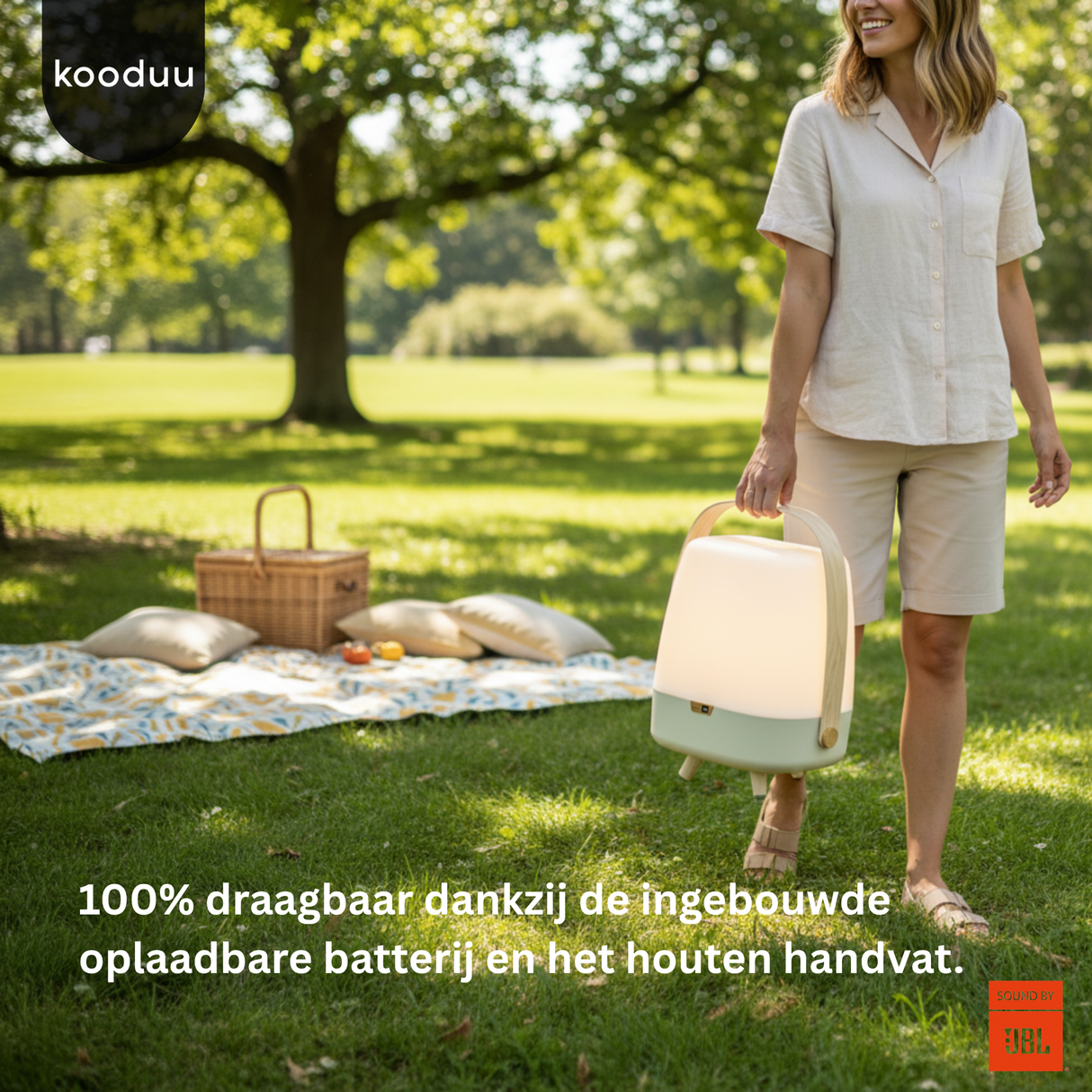 Woman holding a portable lantern in a park with a picnic setup, featuring the brand 'kooduu'.